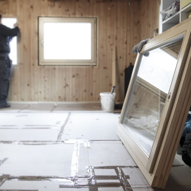 A worker installs new windows in a wooden interior, with energy efficient windows ready for installation in the foreground.