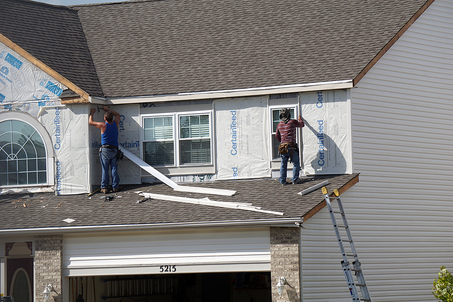 Two workers installing white siding on a house, using ladders and tools to secure the panels over a protective barrier.